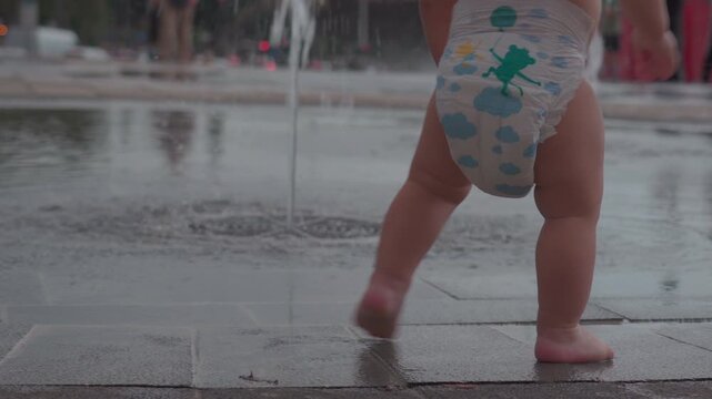 Close-up of toddler in diaper walking barefoot toward water stream of urban fountain, legs and feet visible on wet pavement, playful summer moment