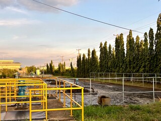 high voltage power lines. An outdoor view of a water treatment or wastewater plant with yellow safety railings, showing a body of churning water, a line of tall trees, and an industrial building under © Jreacher