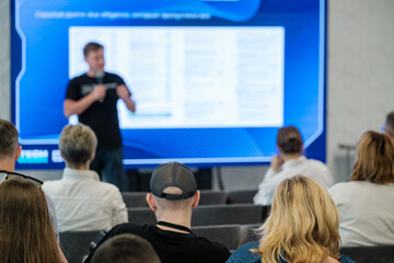 Conference audience attending presentation with speaker in front of informational screen. Modern...