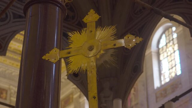 Golden processional cross with radiant sunburst design mounted on ornate column, displayed inside historic basilica with arched windows and decorative ceiling in background