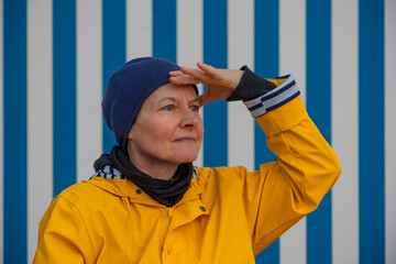 Luc-Sur-Mer, France - 08 07 2025: A mature woman wearing a yellow raincoat and a hat looks at the horizon sitting on a bench with a blue and white striped wall behind at sunrise