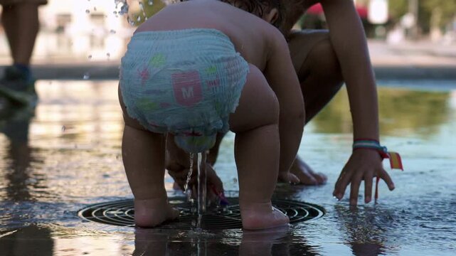 Baby crouching in diaper over water fountain jet with droplets splashing while older sibling bends nearby in playful summer scene at urban public park