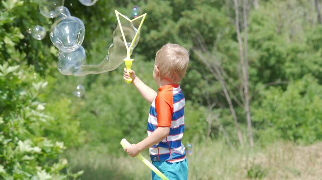 A young boy is making giant bubbles with a yellow bubble wand. He is playing outdoors surrounded by lush greenery. The scene captures the joy and wonder of a sunny day in nature.