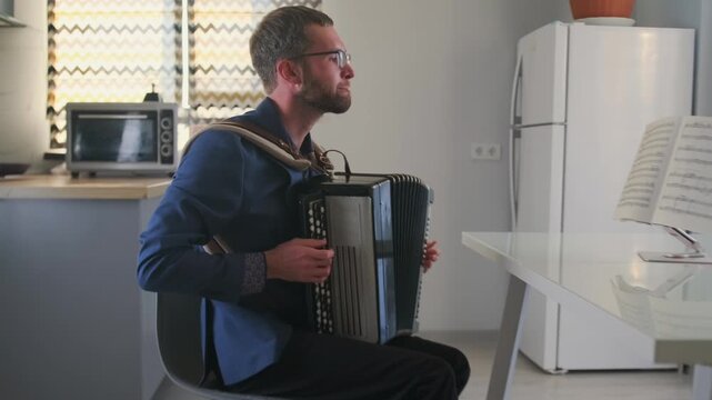 Young man in glasses practices bayan accordion while taking an online music course. Sitting in his kitchen with sheet music on a stand. E-learning, music training, and self-guided lessons.