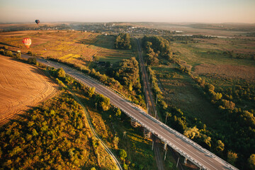 Hot air balloons flying over road, railway, and fields at sunset