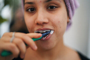 A young woman with a towel on her head brushes her teeth. She uses a green toothbrush with toothpaste foam visible. This captures a daily hygiene routine.