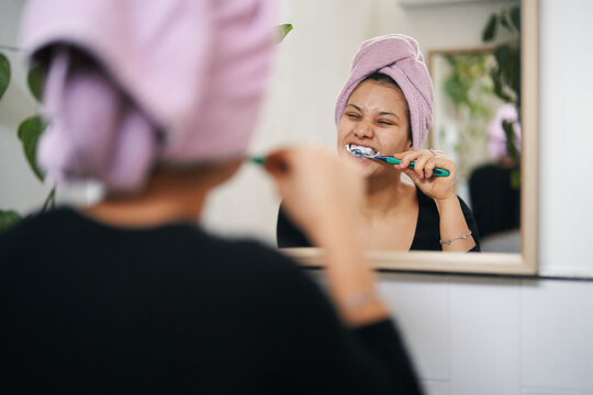 A woman with a towel on her head brushes her teeth in the bathroom mirror. She is focused on her oral hygiene routine, ensuring clean teeth and fresh breath.