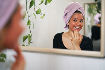 A smiling young woman with a pink towel on her head and a red face mask looks at her reflection in the mirror. She is touching her chin during her skincare routine.