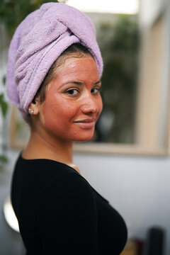 A young woman with a pink towel wrapped around her head has a red face mask applied. She looks over her shoulder, smiling at the camera, enjoying a relaxing beauty treatment.
