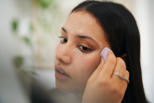 A young woman applies a beauty product to her face with a soft sponge. She is focused on her reflection, engaging in her daily skincare or makeup routine.