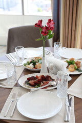 A neatly arranged wooden dining table for four, featuring white plates, crystal glasses, and assorted gourmet dishes