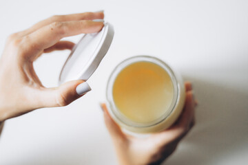 A close-up of a woman hand opening a jar with a glossy, light-colored substance inside, set against a clean, minimalist background.