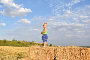 A little boy poses while standing on a pile of straw bales.