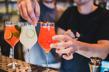 Three colorful cocktails with garnishes on a bar counter, with bartenders preparing and serving drinks in a vibrant, professional mixology setting