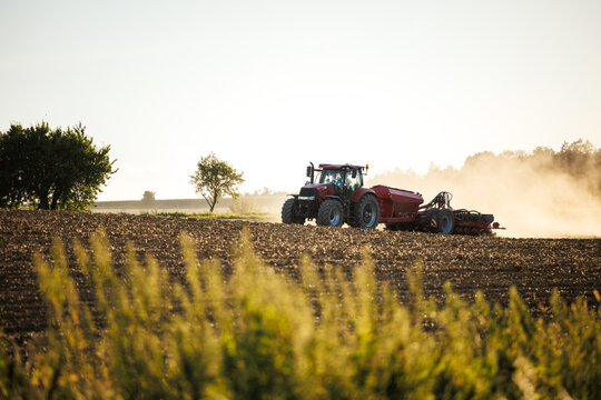 Modern agricultural tractor with seeder on dusty field during summer sowing new crops for next season. Farming machinery preparing soil and planting seeds in rural countryside