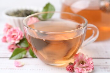 Tasty rose tea in glass cup and flowers on white wooden table, closeup