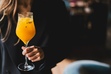 Woman holding a wine glass with vibrant orange cocktail, garnished with ice and passion fruit seeds, against a dark blurred background.