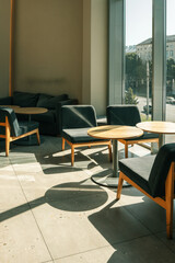 Modern cafe interior with wooden tables, soft chairs, and sunlight casting shadows on the floor near large windows. Cozy urban space with natural light and contemporary furniture design