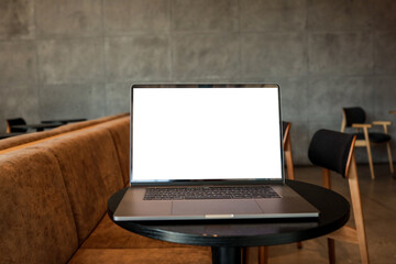 Open laptop with blank white screen on small round table in modern cafe interior with chairs and...