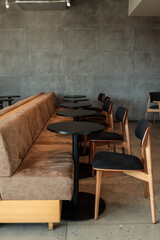 Modern café interior with brown sofa seating, wooden chairs and round black tables against a concrete wall, minimalistic design for restaurant or coworking space