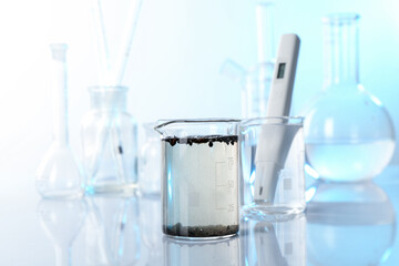 Glass beaker with dirty water, other glassware and digital pH meter on mirror surface