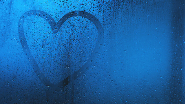 Heart shape is drawn on wet foggy bathroom glass. Romantic and emotional. Droplets water and cool blue tones evoke feelings of love and solitude.