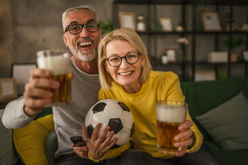 Mature husband and wife watch football match and hold ball and beer