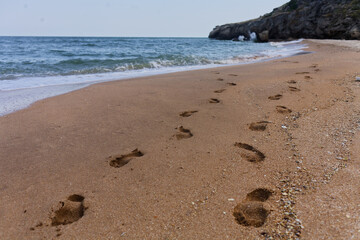Footprints lead towards calm water's edge on sandy beach, gentle waves lap at shore while rocky cliffs stand tall in background. Pebbles scatter near tide line, creating tranquil seaside scene perfect