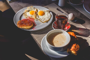 Table is set for a delightful breakfast featuring fried eggs, a pancake with jam, a glass of tea, a bowl of soup, a boiled egg, and a croissant, creating a visually appealing and appetizing scene