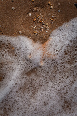 Ocean foam and wet sand glisten under sunlight, scattered seashells and bubbles left behind by receding tide. Bubbles shimmer alongside tiny sand grains and seashells, offering serene beachcombing