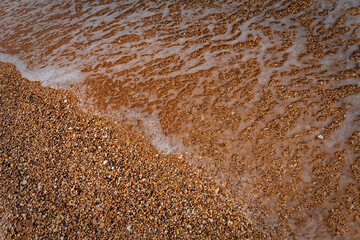 Wet sand displays intricate ripples and small, smooth pebbles near beach edge where shallow water gently laps, creating soft reflections. Top-down aerial view showcases earthy tones and organic