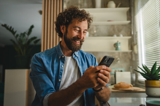 Smiling man using smart phone in kitchen reading good news