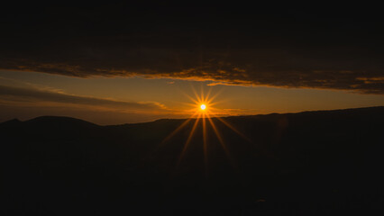 Majestic sunset over distant mountains, with vibrant orange and yellow rays piercing through dark...