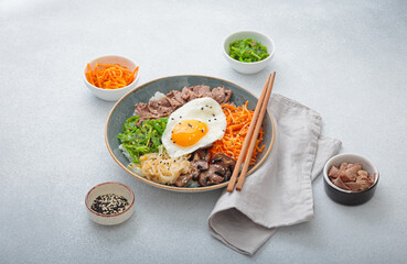 Bibimbap, authentic korean dish with a steamed rice topped with fried egg, beef, colorful vegetables, traditional dish of Korea on light background with chopsticks