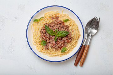 Delicious spaghetti bolognese is garnished with fresh basil leaves and parmesan, served on a white plate next to cutlery on a napkin top view