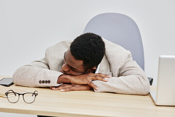 Relaxed black man asleep at office desk wearing beige blazer with closed eyes in a modern workspace...