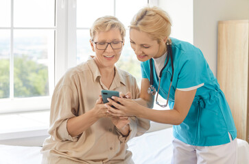 Nurse or caregiver helping elderly woman with usage of smartphone, downloading app for monitoring...