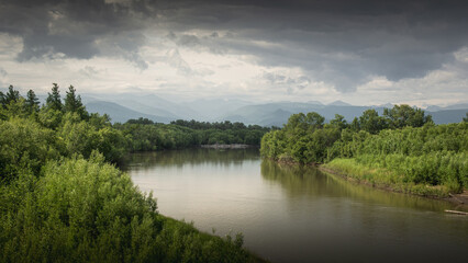 Fototapeta premium Serene river flows through lush green landscape under dramatic cloudy sky, surrounded by distant mountains, creating a peaceful natural scene with vibrant vegetation