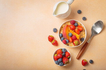 Oatmeal porridge with peach slices, strawberries, blueberries, raspberries and milk jug on beige table, concept of healthy and delicious breakfast, copy space
