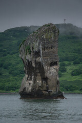 Majestic rock formation rises dramatically from calm waters, surrounded by lush green hills under a...