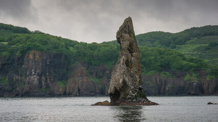 Majestic rock formation rising from calm waters, surrounded by lush green hills under a cloudy sky,...