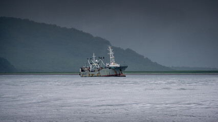 Fishing vessel navigating calm waters under a moody sky, surrounded by lush green hills, showcasing...