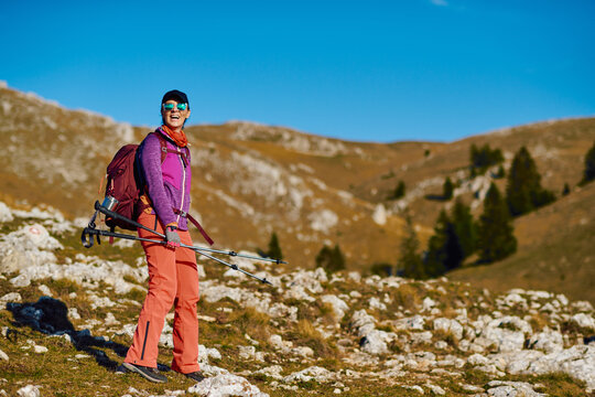 Portrait of young female hiker wearing colorful outfit and sunglasses holding trekking poles in mountain landscape