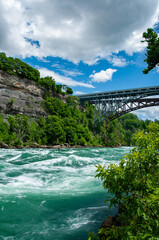 The powerful, churning turquoise rapids of the Niagara River, flowing through a lush green gorge. The large Whirlpool Rapids steel bridge spans the cliffs in the background.