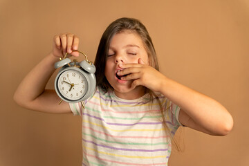 Back to school. 9 year old girl yawning and looking tired while holding gray alarm clock showing time to go to school on beige background. Time management concept.