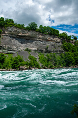 The powerful, turquoise rapids of the Niagara River flowing through a lush green gorge with rocky cliffs. The sky is partly cloudy, and the trees are vibrant green, suggesting a warm and sunny day.