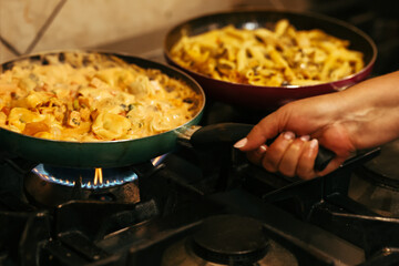 Close-up of homemade creamy pasta being cooked in a pan on a gas stove, with a person holding the handle. Warm and authentic kitchen atmosphere