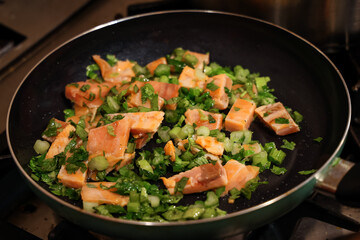 Fresh salmon cubes with green vegetables and herbs being cooked in a frying pan. Healthy homemade meal preparation with colorful ingredients.