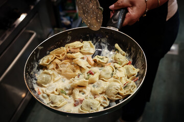 Chef holding a skillet of creamy tortellini with vegetables, stirring on the stove. Close-up of homemade filled pasta cooking in a warm kitchen atmosphere
