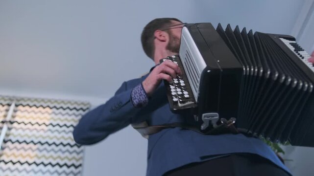 Dramatic low-angle close-up of a young bard and singer-songwriter emotionally performing on the accordion near a sheet music stand, during a private music session.